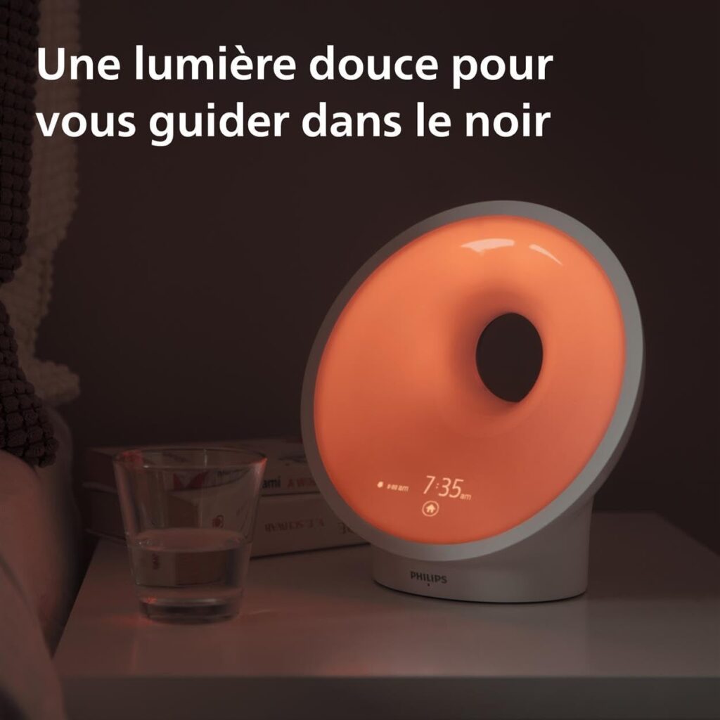 Le radio-réveil Philips SmartSleep sur une table de chevet, avec une lumière tamisée rouge orangé. Un verre d'eau est à côté....
