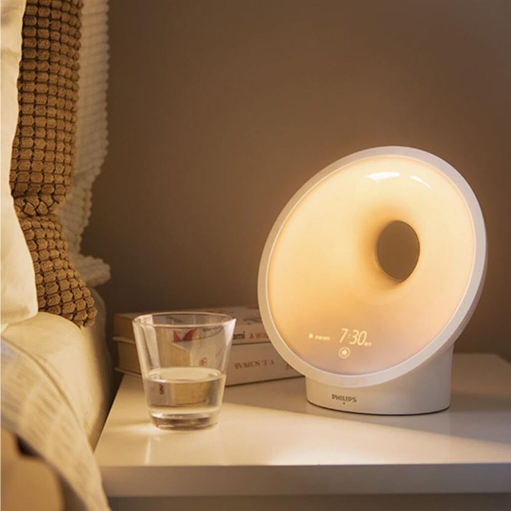 Le radio-réveil Philips SmartSleep, posé sur une table de chevet, avec la lumière allumée. Un verre d'eau est à côté.