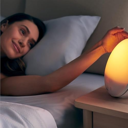 Une femme au lit tend la main vers le radio-réveil Philips SmartSleep posé sur une table de chevet. Le réveil est allumé et i...