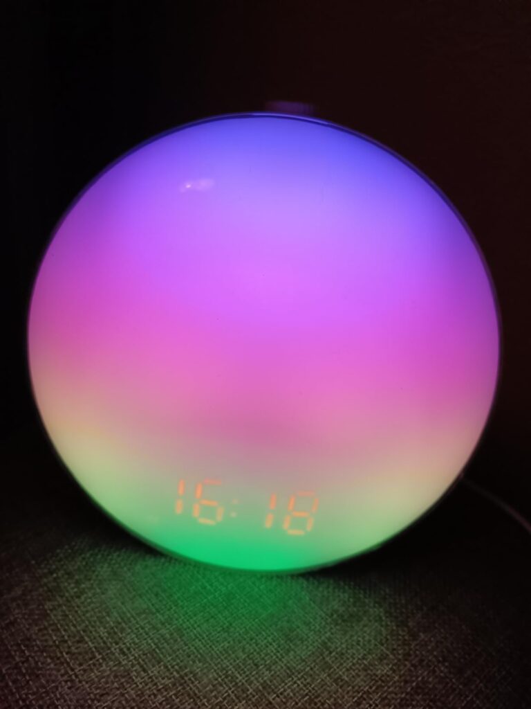Le réveil est posé sur une table de chevet, allumé avec une lumière multicolore et l'heure affichée.