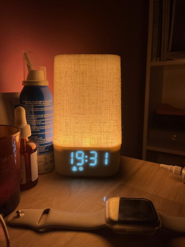 Appareil allumé sur une table de chevet, affichant 19:31. On voit un flacon, une montre connectée et une étagère en arrière-p...