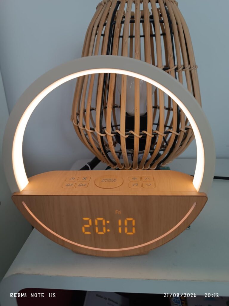 Le réveil-lampe de luminothérapie est posé sur une table de chevet, allumé. L'heure affichée est 20:10. On voit une partie d'...