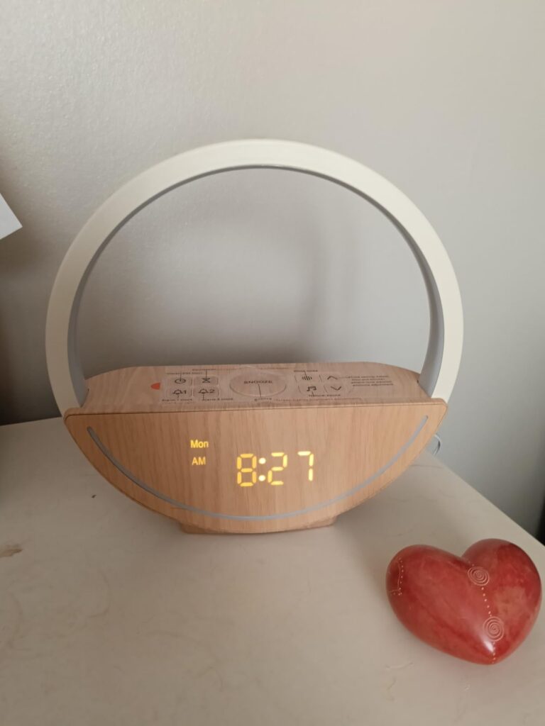 Le réveil-lampe est posé sur une table de chevet, allumé. L'heure affichée est 8:27. À côté du réveil se trouve un petit obje...