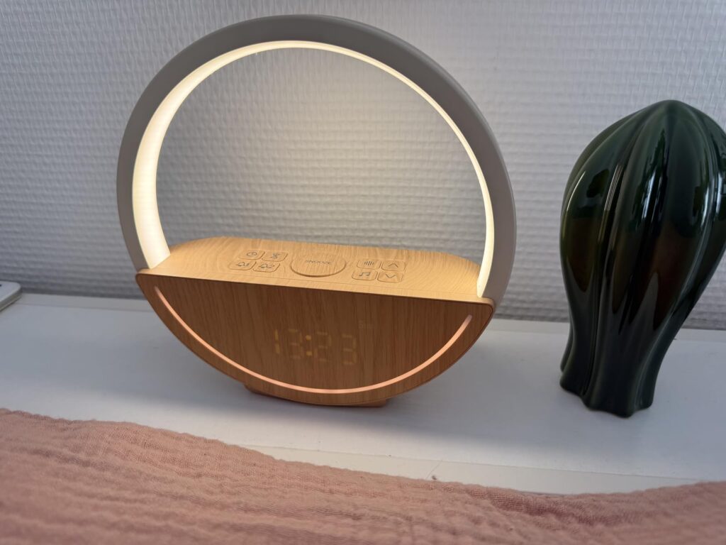 Le réveil-lampe est posé sur une table de chevet, allumé. L'objet est montré sous un angle différent des images précédentes.