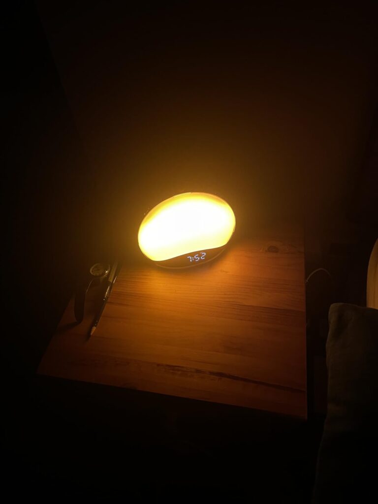 Le réveil lumière de face, allumé en lumière orange, posé sur une table en bois. La lumière est tamisée. Le réveil est ovale....