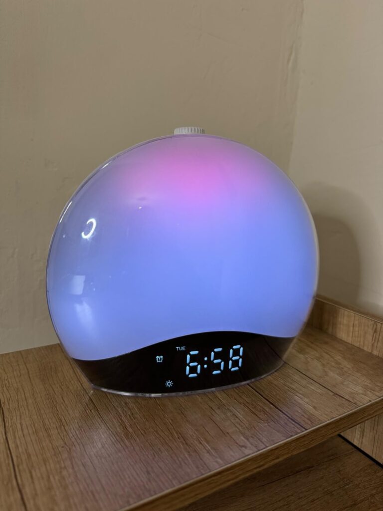 Le réveil lumière de face, allumé en lumière multicolore, posé sur un meuble en bois clair. L'heure est affichée en numérique...