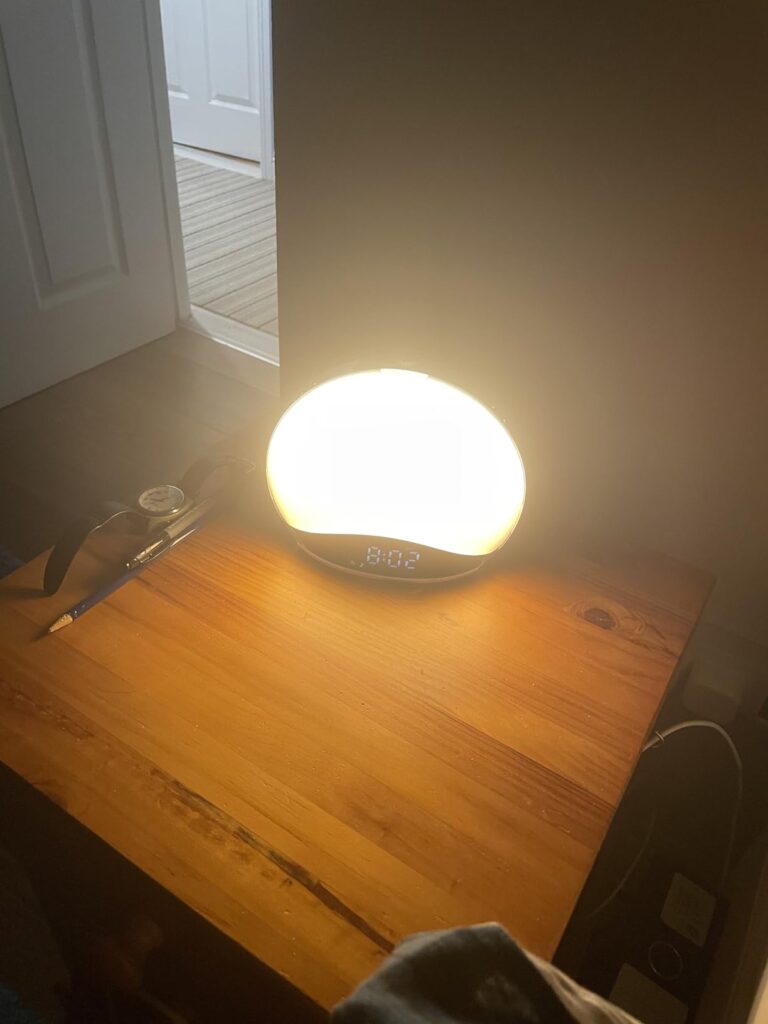 Le réveil lumière de face, allumé en lumière blanche vive, posé sur une table en bois. Le réveil est ovale, et son contour es...