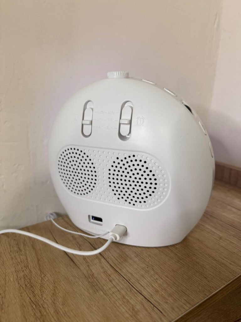 Le réveil lumière de dos, couleur blanche, posé sur un meuble en bois clair. Des hauts parleurs sont visibles. Un port USB es...