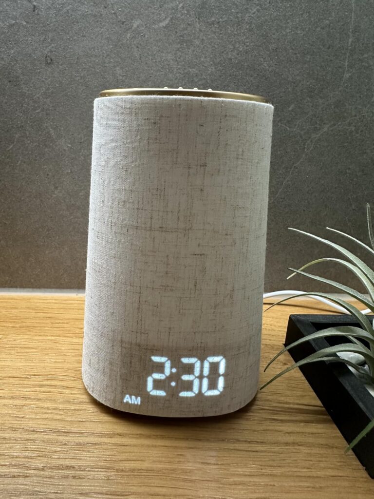 Le produit est posé sur une table en bois, affichant l'heure 2:30 AM, avec une plante en arrière-plan.