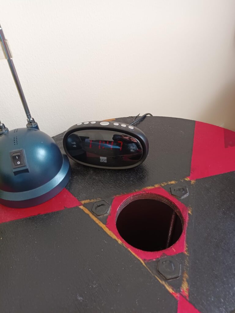 Vue oblique sur le radio-réveil posé sur une table, avec une lampe et un trou dans la table.