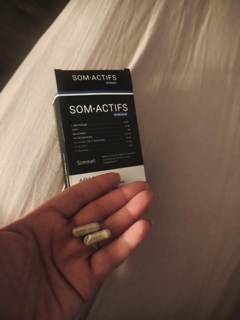 Boîte de SOM-ACTIFS et deux gélules dans une main sur un lit.