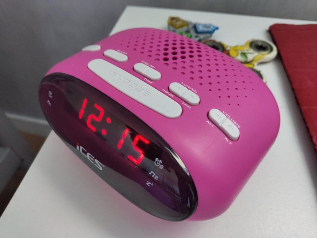 Radio-réveil rose en situation sur une table de chevet, affichant l'heure 12:15.
