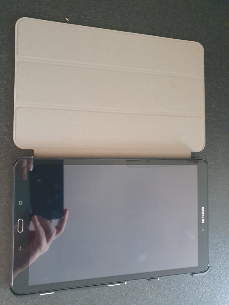 Tablette avec une coque de couleur claire, ouverte et pos'ee sur une surface fonc'ee. L'ecran est 'eteint et l'on voit un ref...