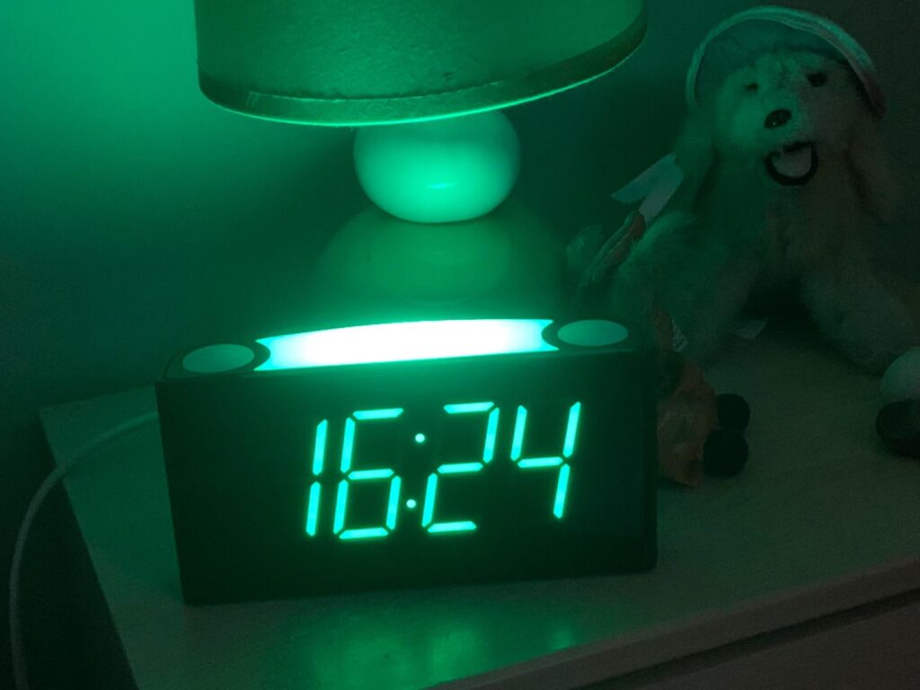 Réveil posé sur un meuble de chevet, l'heure est en vert. On perçoit une lampe de chevet et une peluche en arrière-plan.