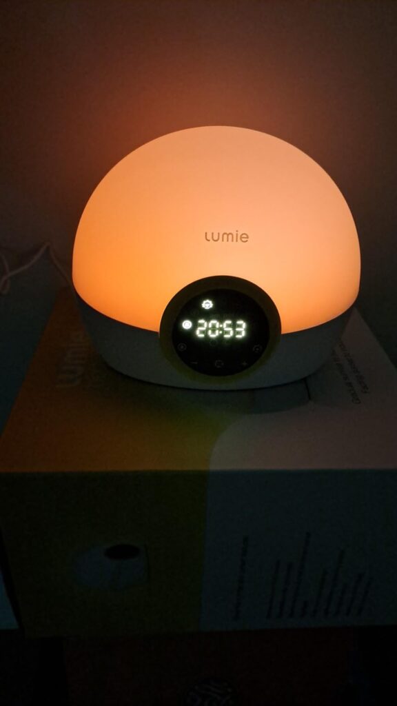 Le réveil Lumie 300 allumé avec une lumière orangée, affichant 20:53, posé sur une table de chevet.