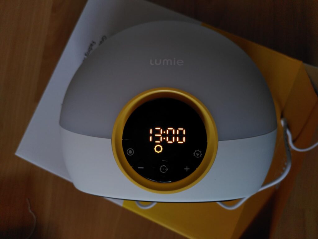Gros plan sur le réveil Lumie 300 posé sur sa boîte, avec l'heure 13:00 affichée et les boutons de contrôle visibles.