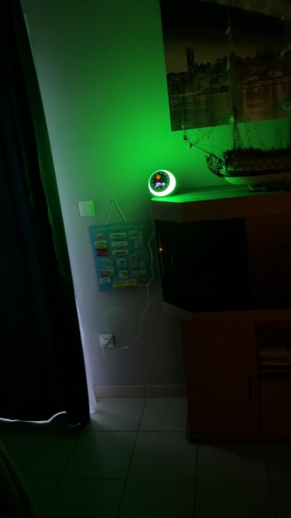Le réveil sur une étagère dans une pièce sombre, avec un éclairage vert projeté sur le mur. Le visuel montre le produit en si...