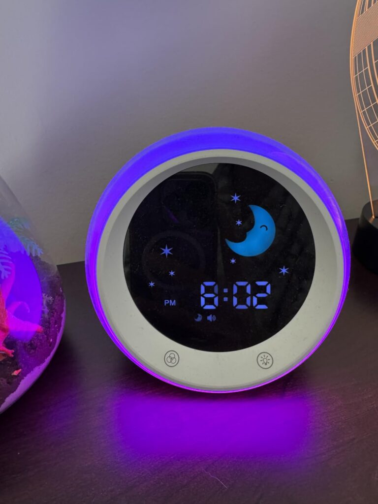 Le réveil sur une table de nuit, dans la pénombre, avec un éclairage violet. Le visuel montre le produit en situation, avec d...