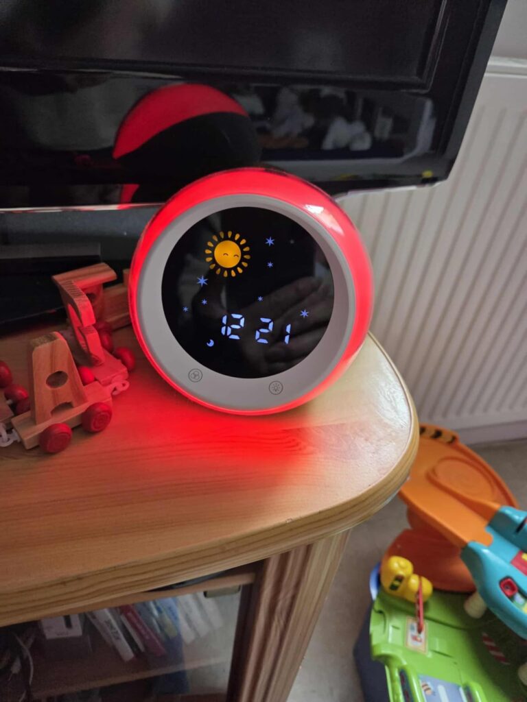 Le réveil sur une table de nuit, dans la pénombre, avec un éclairage rouge. Le visuel montre le produit en situation, avec de...