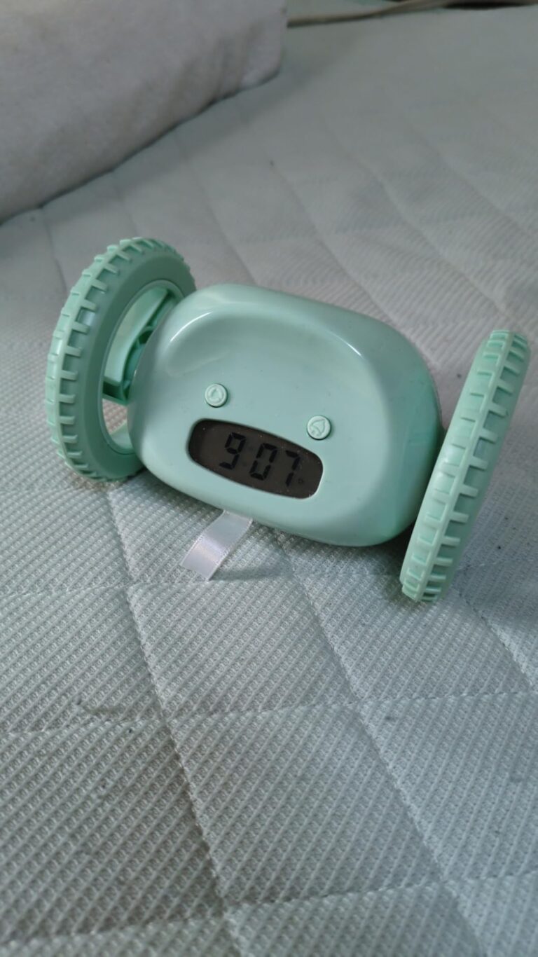Réveil à roulettes vert affichant 9:07, posé sur un lit avec un drap blanc matelassé.