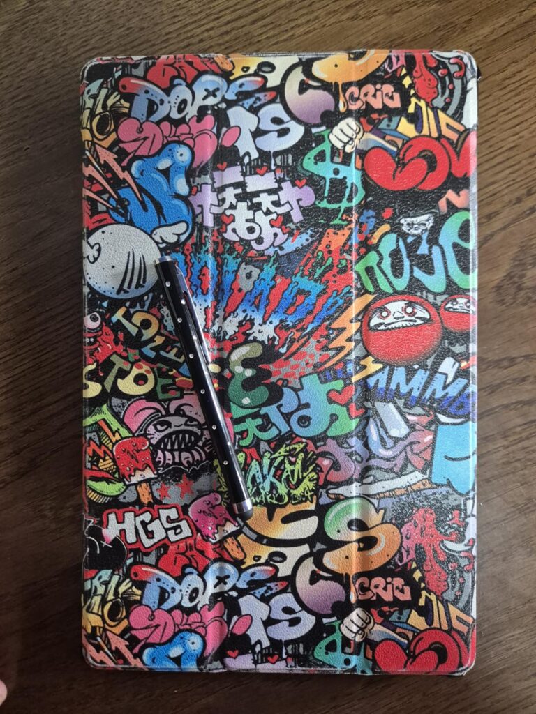 Étui de tablette avec motif graffiti, un stylo noir est posé dessus. C'est le même modèle que le visuel 1, mais sur une table...