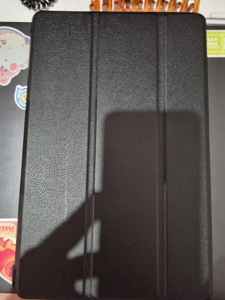 La tablette est présentée dans son étui fermé, vue de face, posée sur une table sombre avec des autocollants. Une ombre cache...