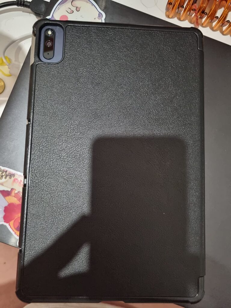 La tablette est présentée dans son étui fermé, vue de dos, posée sur une table sombre avec des autocollants. Une ombre cache...