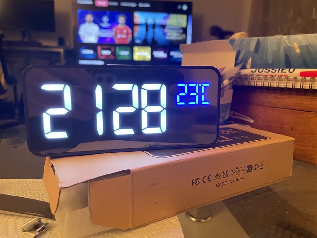 Le réveil allumé, posé sur sa boîte d'emballage ouverte. Une télévision est visible à l'arrière-plan.