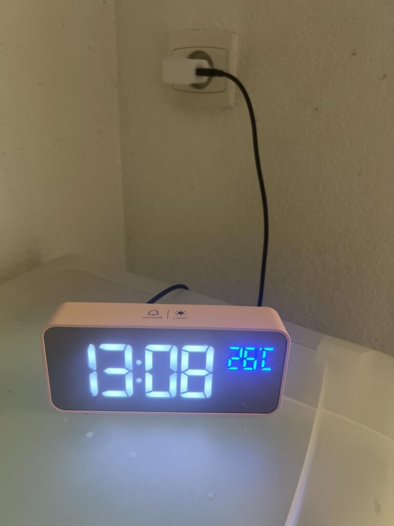 Le réveil est branché sur une prise murale, affichant l'heure 13:08 et la température 26°C, dans un environnement clair.
