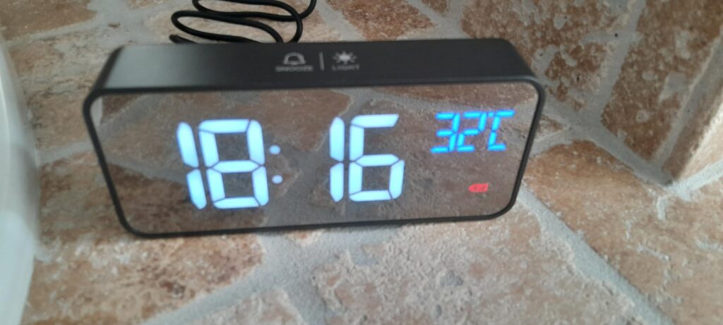 Le réveil sur une surface carrelée, affichant l'heure 18:16 et la température 32°C. La façade est réfléchissante.