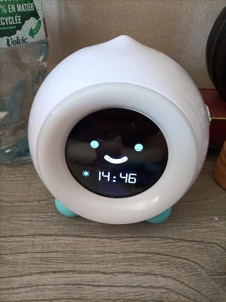 Le réveil sur une table en bois, avec une bouteille en plastique recyclé derrière. L'heure affichée est 14:46.