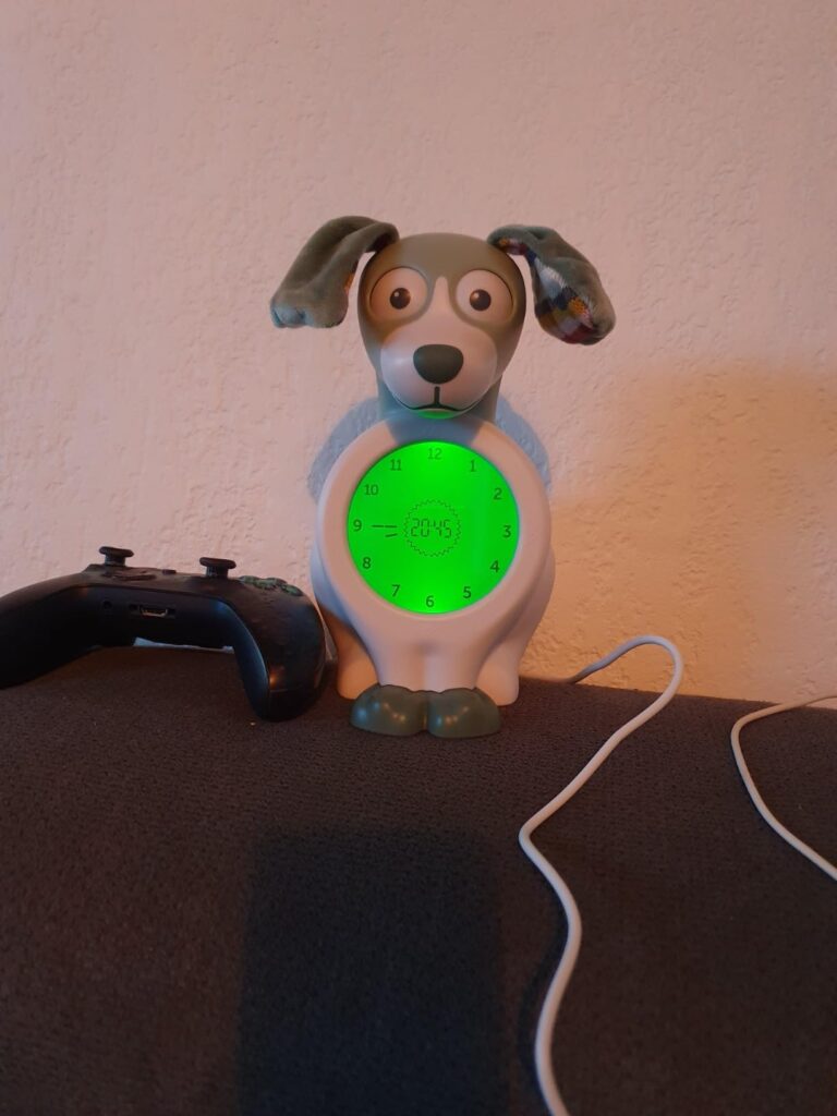 Réveil chien Zazu posé sur un meuble, avec une manette de console de jeu à côté, le cadran est allumé et vert.