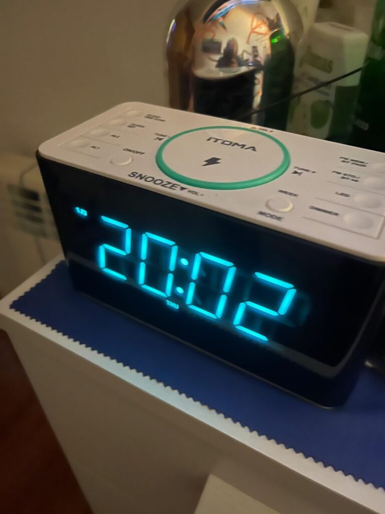 Radio-réveil en situation sur une table de nuit, vue de face. La lumière d'ambiance bleue sous le réveil est allumée. L'affic...