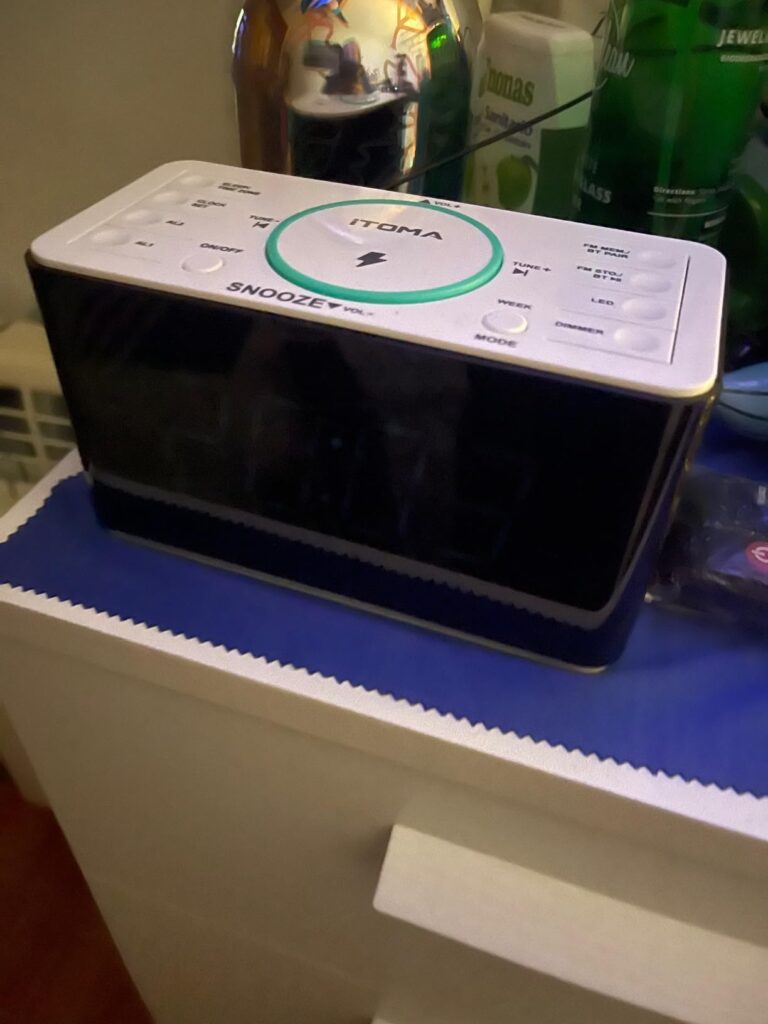 Radio-réveil en situation sur une table de nuit, vue de dessus. La lumière d'ambiance est éteinte. Le dessus du réveil est bl...