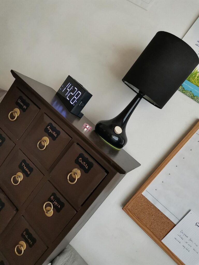 Réveil posé sur une commode à tiroirs, à côté d'une lampe. L'heure affichée est 14:28.