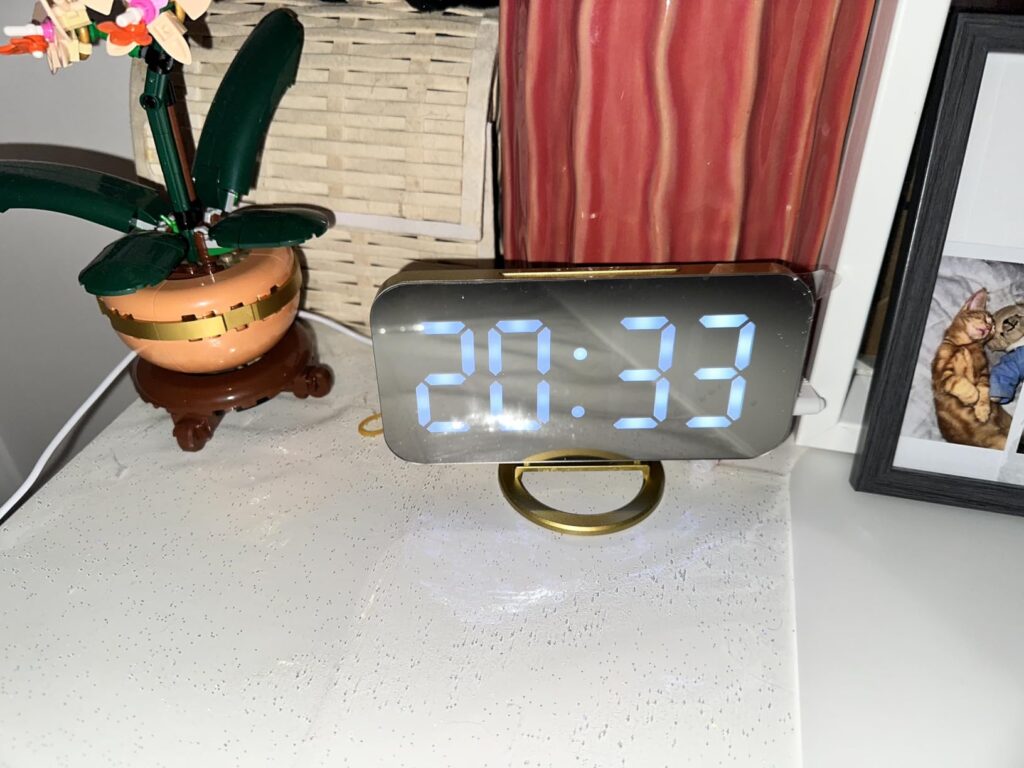 Le réveil est posé sur une table de chevet, affichant 20h33. Des objets décoratifs sont autour.