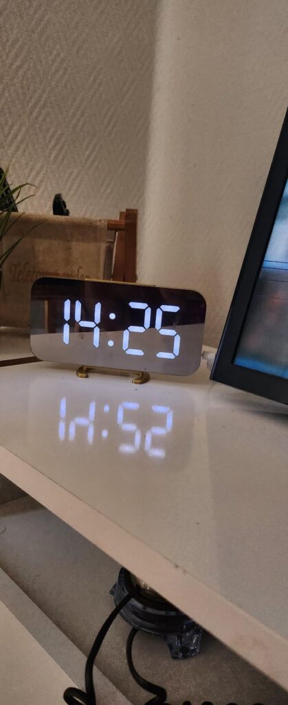 Le réveil est posé sur un meuble, affichant 14h25, avec le reflet de l'heure sur la surface.