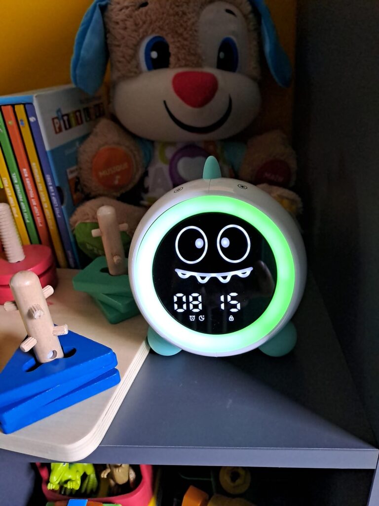 Le réveil est placé sur une étagère à côté d'un jouet en peluche et d'autres objets, affichant l'heure 08:15 avec une express...