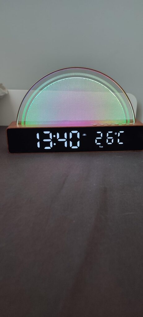 Le réveil est posé sur une surface sombre, l'écran affiche '13:40' et '26°C'. La lumière est colorée et diffusée vers le haut...