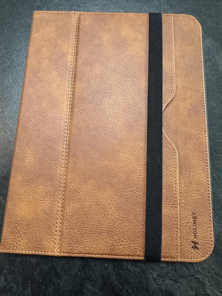Vue de dessus d'une coque pour tablette en simili-cuir brun, de face, fermée par un élastique noir. Le logo de la marque est...