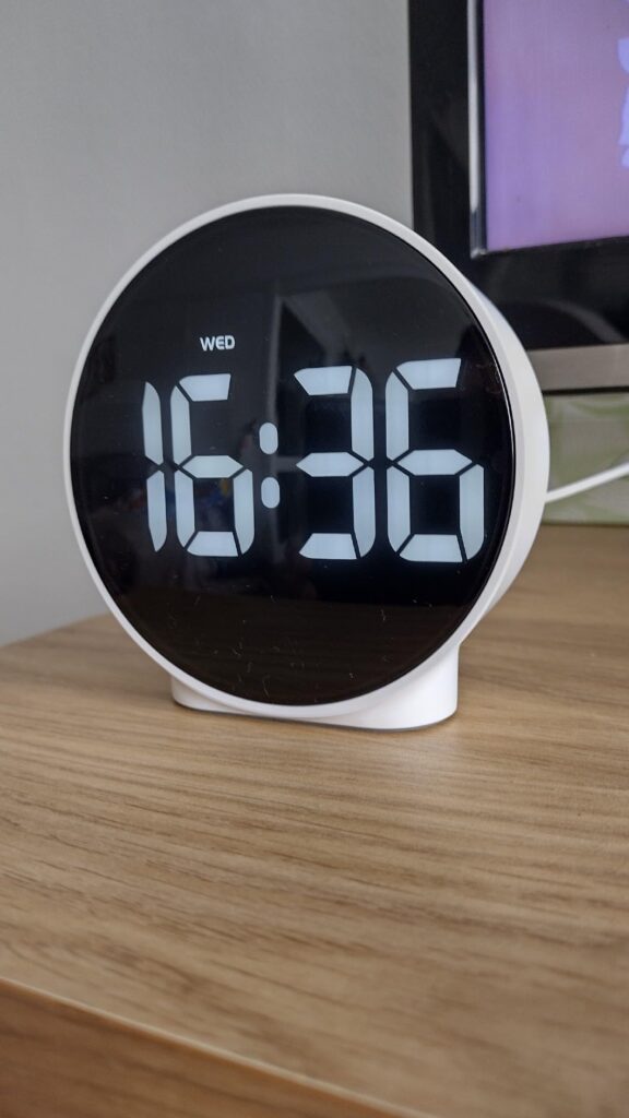 Le réveil sur un meuble en bois, l'heure affichée est 16h36. L'angle de vue est légèrement différent du visuel 0.