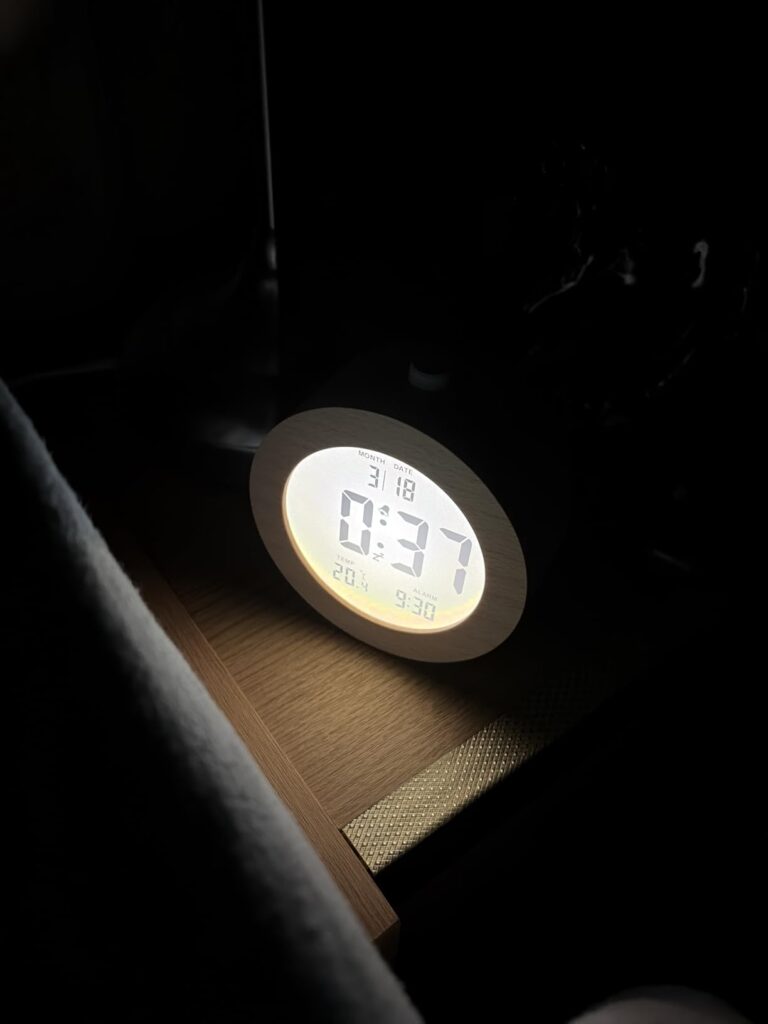 Un réveil numérique à cadre en bois de forme ronde est posé sur une surface dans l'obscurité, l'écran lumineux affichant l'he...