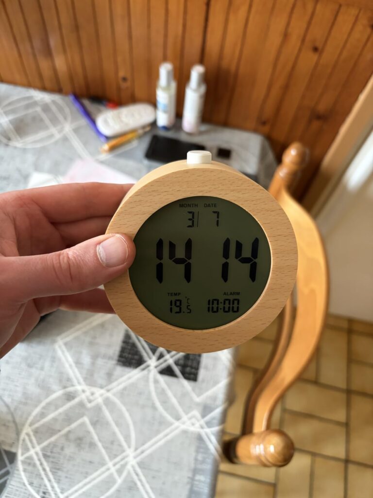 Un réveil numérique à cadre en bois de forme ronde est tenu dans une main, affichant l'heure, la date, la température et l'he...