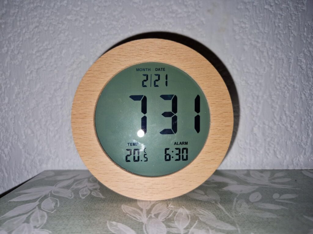Un réveil numérique à cadre en bois de forme ronde, vu de face, avec l'écran affichant la date, l'heure, la température et l'...