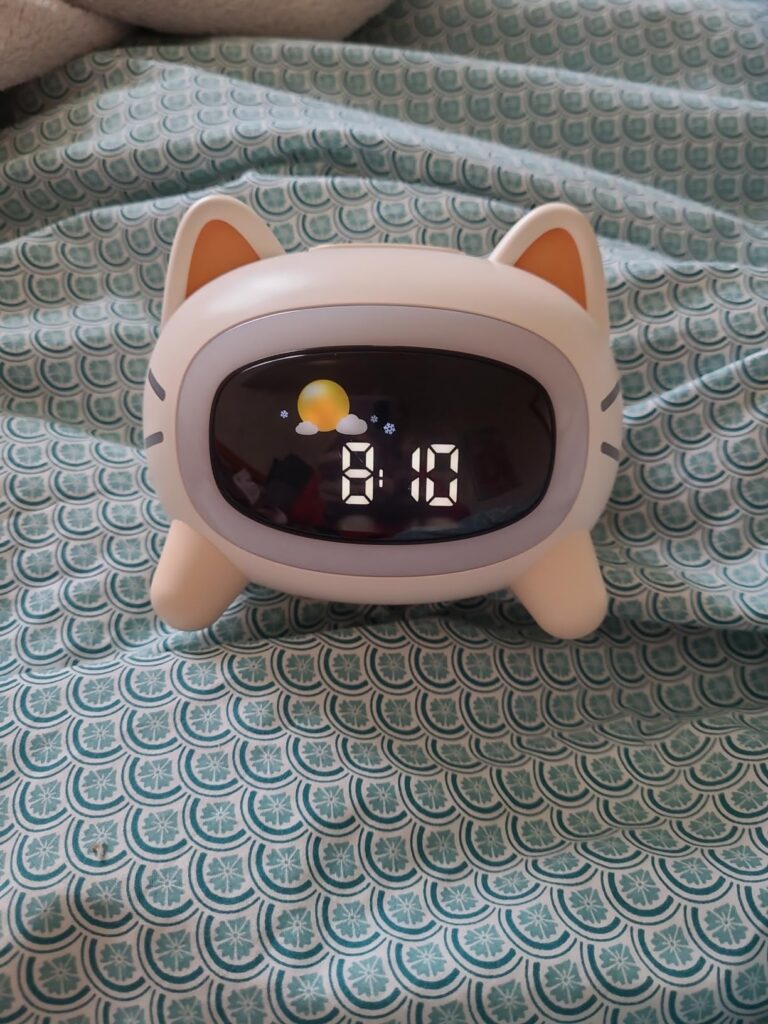 Réveil chat posé sur un lit avec l'heure 8:10 et un affichage du soleil et d'un nuage, sans éclairage d'ambiance.