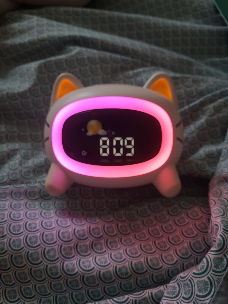 Réveil chat posé sur un lit avec un éclairage rose et l'heure 8:09. L'affichage indique également un soleil et un nuage.