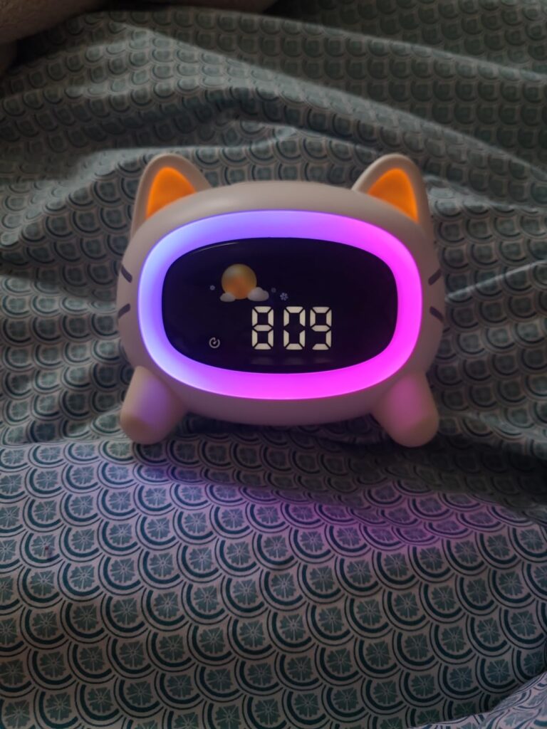 Réveil chat posé sur un lit avec un éclairage multicolore (rose et violet) et l'heure 8:09. L'affichage indique également un...