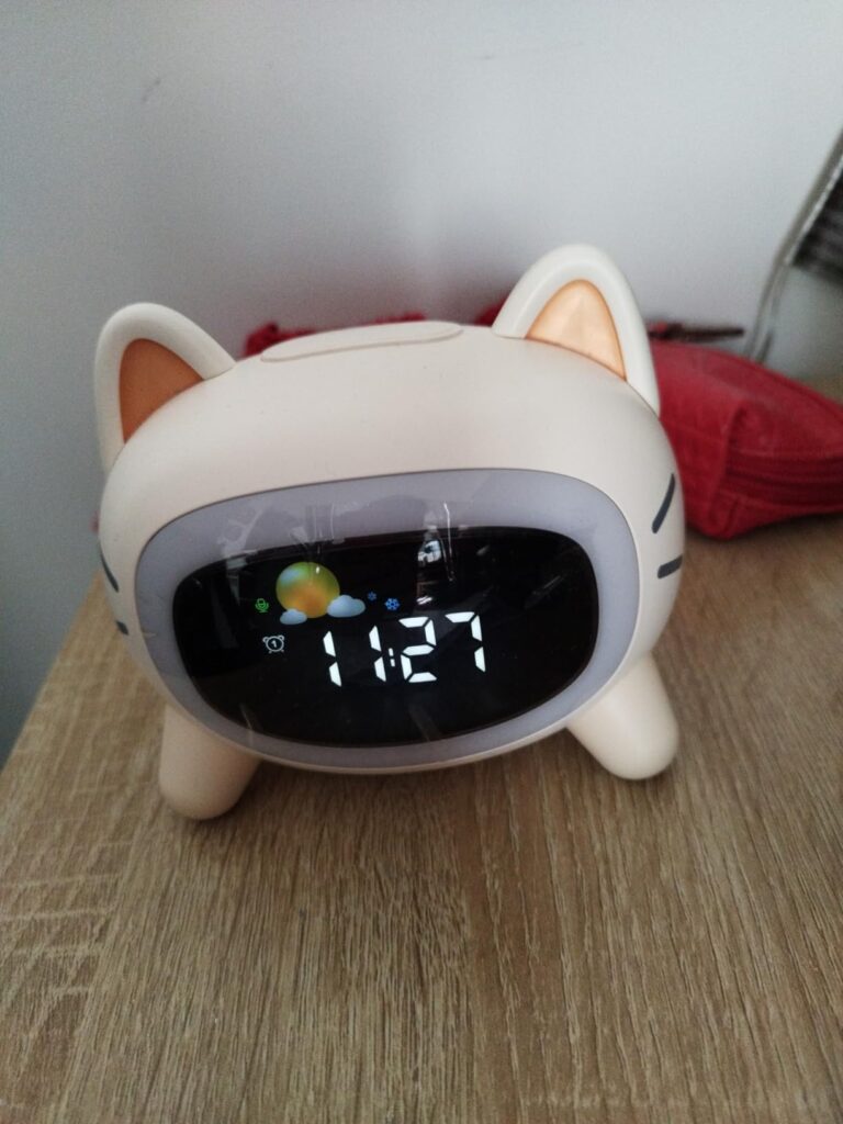 Réveil chat posé sur une table de chevet avec l'heure 11:27 et un affichage du soleil et d'un nuage.