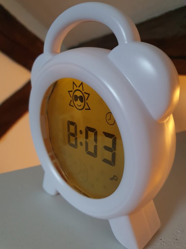 Le réveil blanc affichant 8h03 et un soleil avec des lunettes de soleil, posé sur une table de chevet dans une chambre faible...