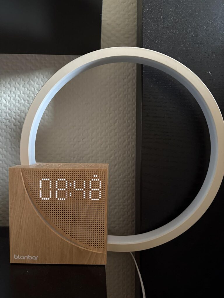 Le réveil-lampe est posé sur une table de chevet en bois clair, allumé, dans une chambre. On y voit l'heure 08:48.